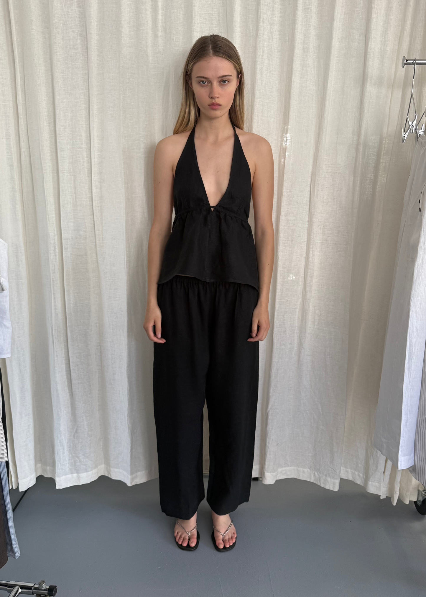 A woman with long, straight hair stands before cream curtains, wearing Deiji Studios’ Linen Button Halter - Black with a deep V-neck and loose black pants. She completes the look with black sandals.