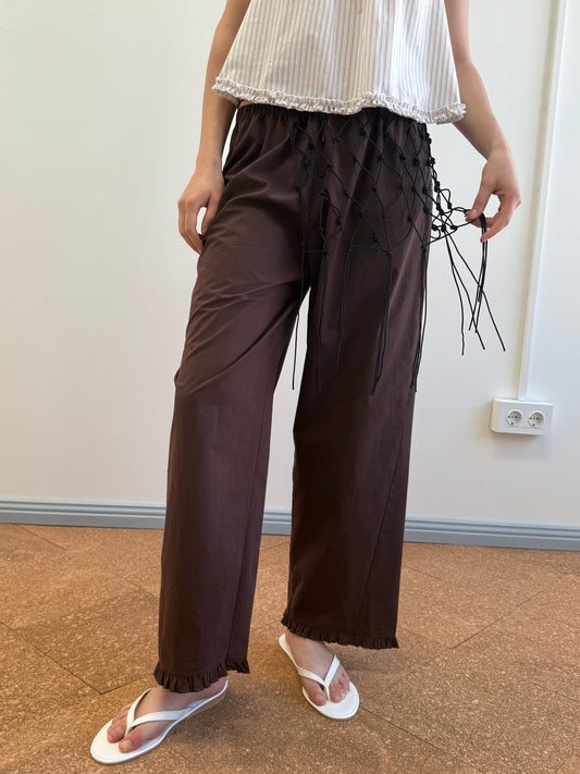 A person stands indoors on a brown floor near a white wall with a power outlet, wearing the Flounce Pant - Umber featuring black string waist details, paired with a white sleeveless top and white flip-flops.
