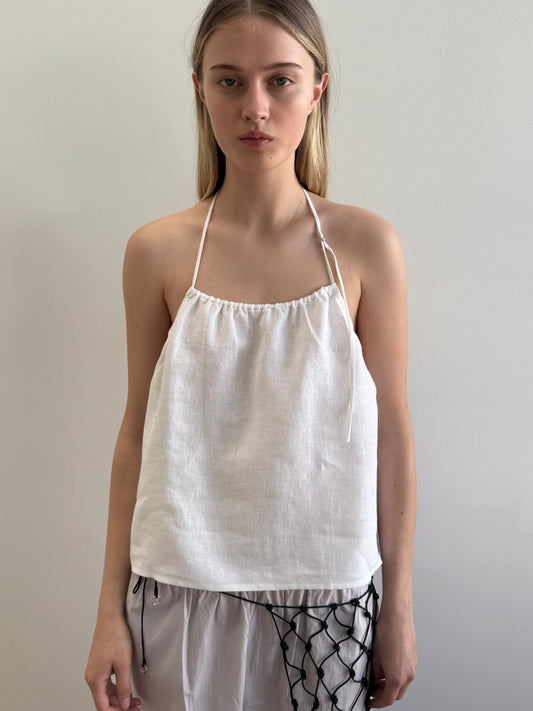 Model wearing the O Ring Halter - White by Deiji Studios, a white linen top with thin tie straps and a gathered neckline, paired with white trousers featuring black macramé rope details.