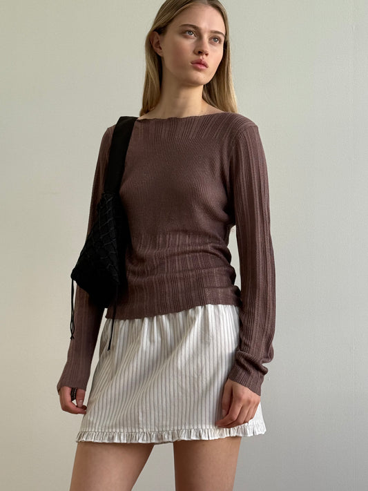 A woman stands against a plain light-colored wall, wearing a ribbed brown long-sleeve top, the Flounce Mini - Gull Stripe with subtle stripes and ruffled hem, and a black shoulder bag. She gazes slightly to the side with a neutral expression.