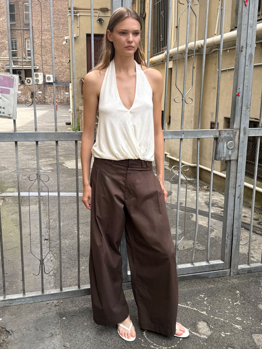 A woman stands outside before a metal gate, wearing a sleeveless white halter top and the Tab Pant - Dark Brown. She has straight hair and a neutral expression, with paved ground and buildings visible behind her.