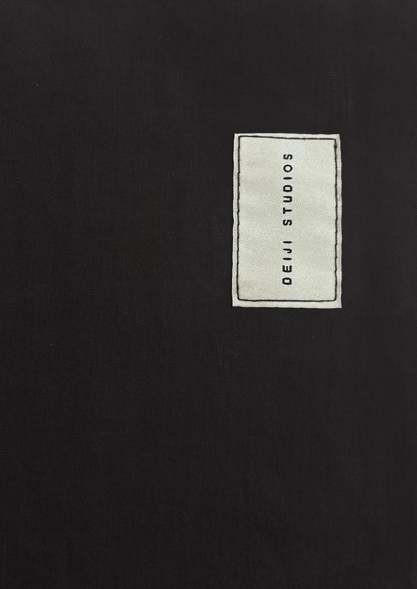 A white rectangular fabric label with black Deiji Studios text is sewn onto the plain black organic cotton Seam Tote.