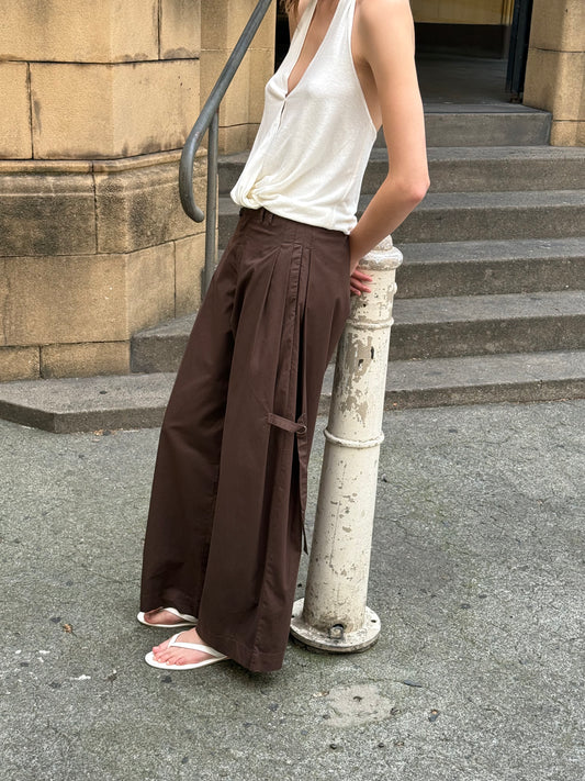 A person stands outdoors by a white post in a sleeveless white top, the Tab Pant - Dark Brown, and white flip-flops. Stone steps and a tan brick building appear in the background.