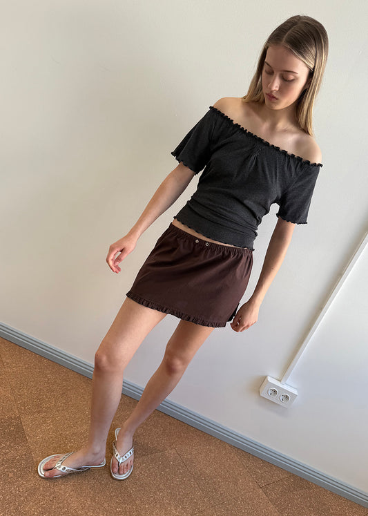 A young woman stands indoors against a light wall, wearing an off-the-shoulder black top, the Flounce Mini - Umber skirt with an elastic waistband, and white sandals. She looks down, arms relaxed at her sides.