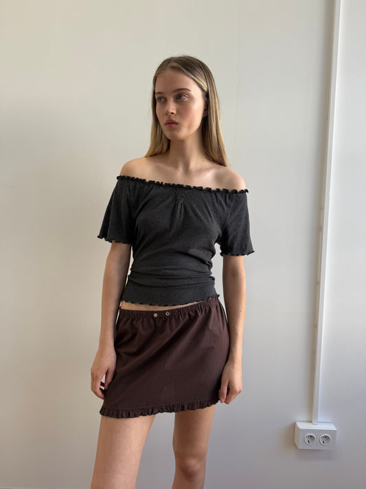 A woman with long, straight hair stands against a plain white wall, wearing the Flounce Tee - Grey and a brown skirt. She looks slightly to the side with a neutral expression.