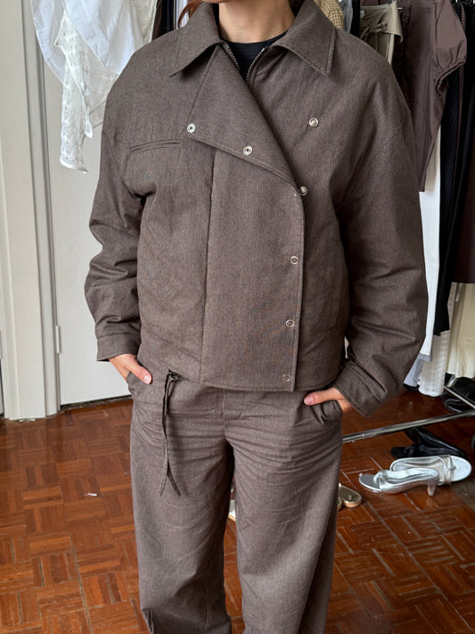A person wearing the Lapel Jacket - Brown Stripe with matching pants stands indoors on a wooden floor, hands in pockets. Clothing items from Deiji Studios appear in the background.