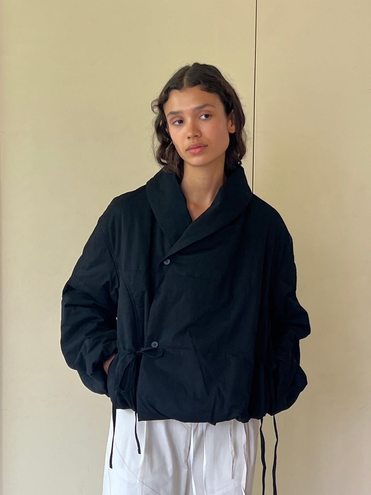 A person with wavy dark hair wears the 0 Bubble Coat - Black, crafted from recycled polyester, paired with white pants against a plain beige background.