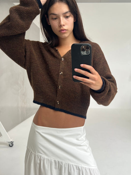 A woman with long dark hair poses for a mirror selfie in a minimal, bright room. She is wearing the Cardi - Tawny textured knit cardigan and a white skirt, holding her phone in one hand against neutral walls.