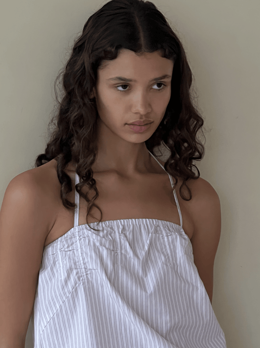 A young woman with long, wavy dark hair stands against a neutral background, wearing the Tube Top - Musk Stripe from Deiji Studios in white with pinstripes, crafted from organic cotton, and looks thoughtfully to the side.