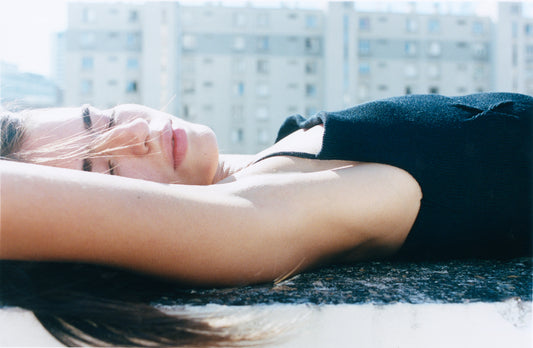 A person in the Fine Knot Tank - Black lies outdoors on their back, eyes closed and sunlit, with tall apartment buildings blurred in the background—Deiji Studios style.