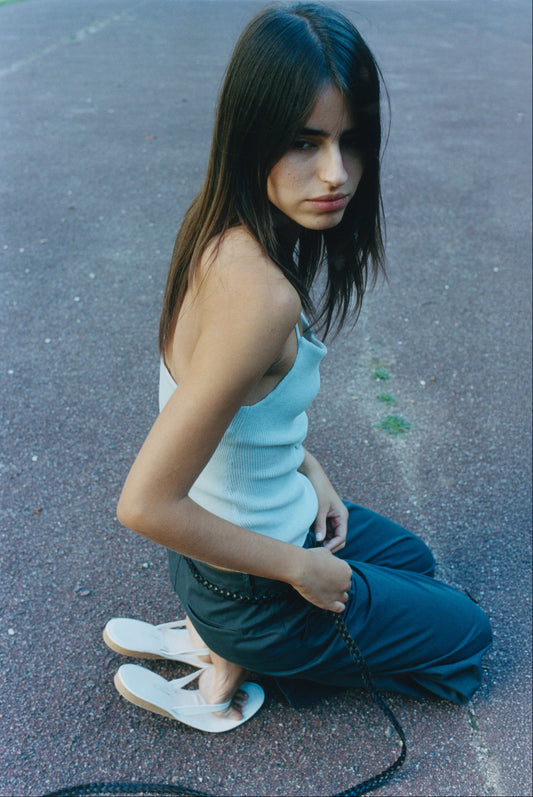 A young woman with long brown hair kneels outdoors, wearing the Fine Knot Tank - Ice Blue by Deiji Studios, dark pants, and white sandals. She gazes calmly to the side, holding a strap in her hand.