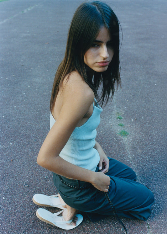 A young woman with long straight hair kneels on a paved surface, wearing a light blue tank top, Zip Pant - Charcoal by Deiji Studios, and white sandals. She gazes pensively to the side.