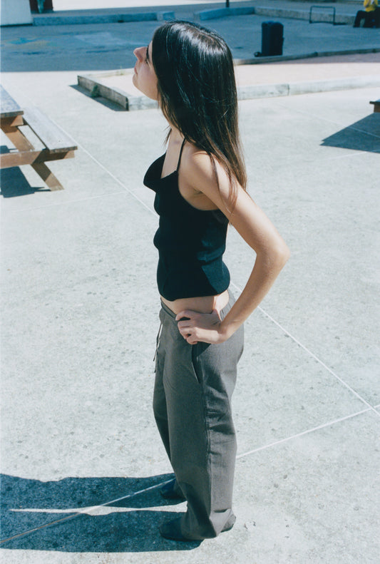 A woman with long brown hair stands outdoors on concrete, wearing a black sleeveless top and the Double Belted Pant - Brown Stripe by Deiji Studios, hands on hips and looking upward into the sunlight near a wooden bench and shadows.