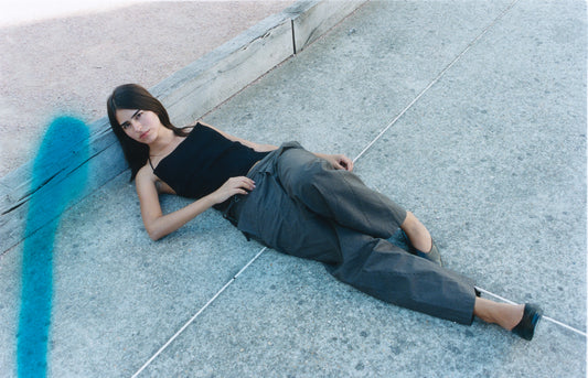 A woman with long dark hair in a black tank top and Double Belted Pant - Brown Stripe by Deiji Studios reclines on a concrete surface outdoors, leaning against a wooden beam. A blue streak is visible on the left side of the image.