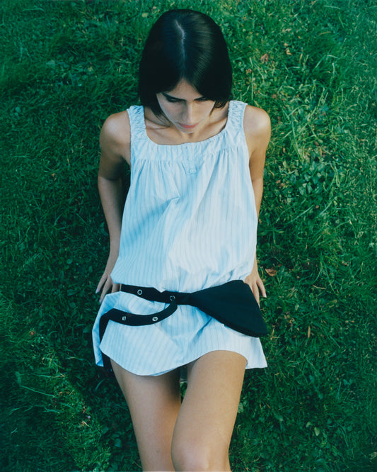 A woman with straight dark hair, wearing a light-colored sleeveless Deiji Studios dress and the Snap Belt Bag - Black—an everyday accessory—sits on green grass, looking down with her hands resting behind her.