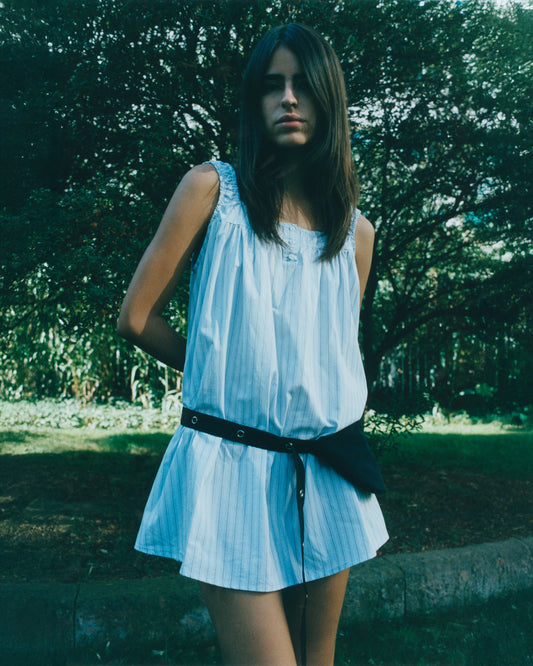 A young woman with long dark hair stands outdoors in a sleeveless white striped mini dress, accessorized with the Snap Belt Bag - Black, posed against a lush green garden—a chic accessory that completes her look.