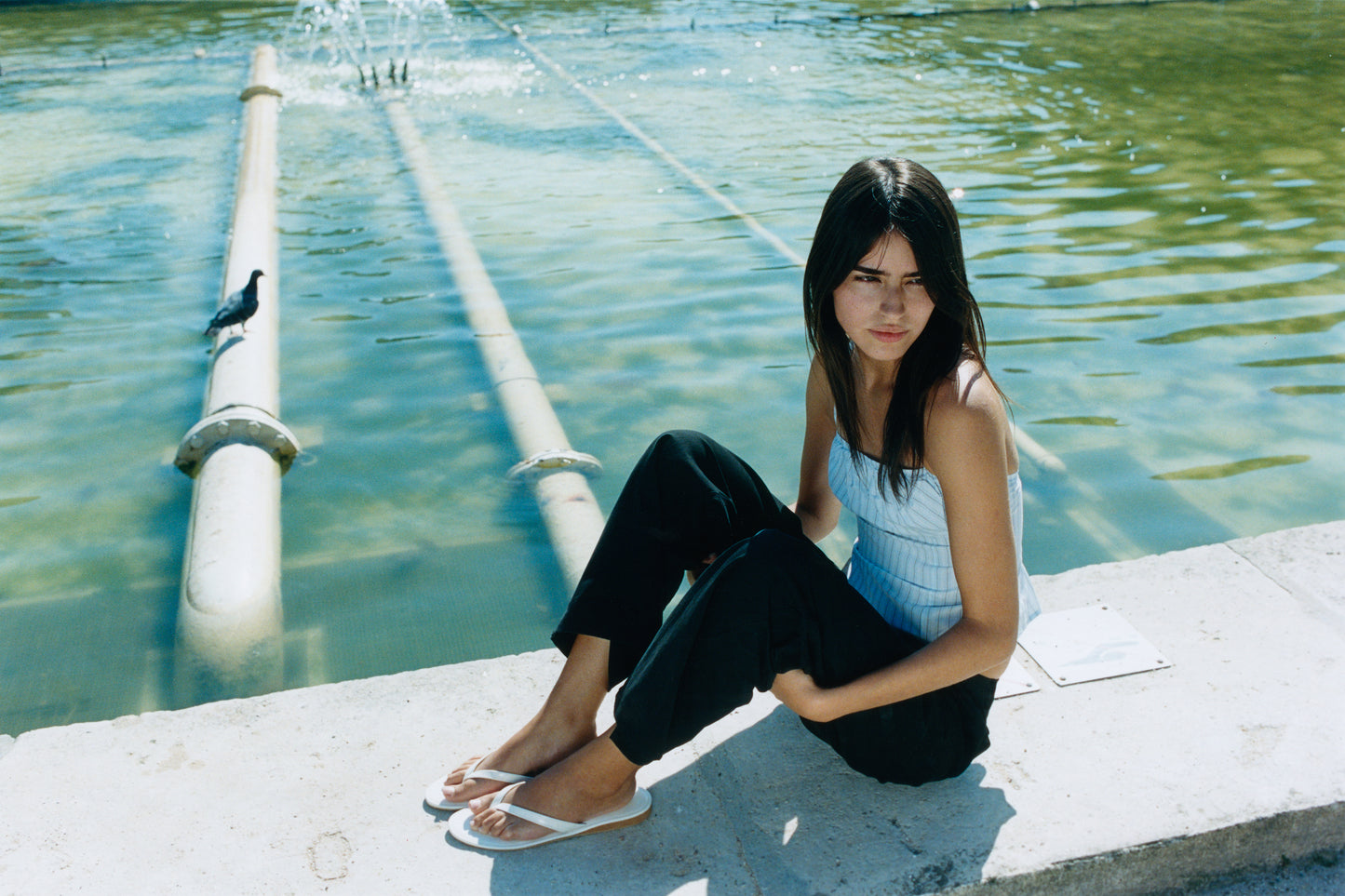 A young woman wearing a light blue tank top and the Zip Pant - Black sits on the edge of a fountain, with clear water and pipes behind her. A pigeon stands on one of the pipes in the background.