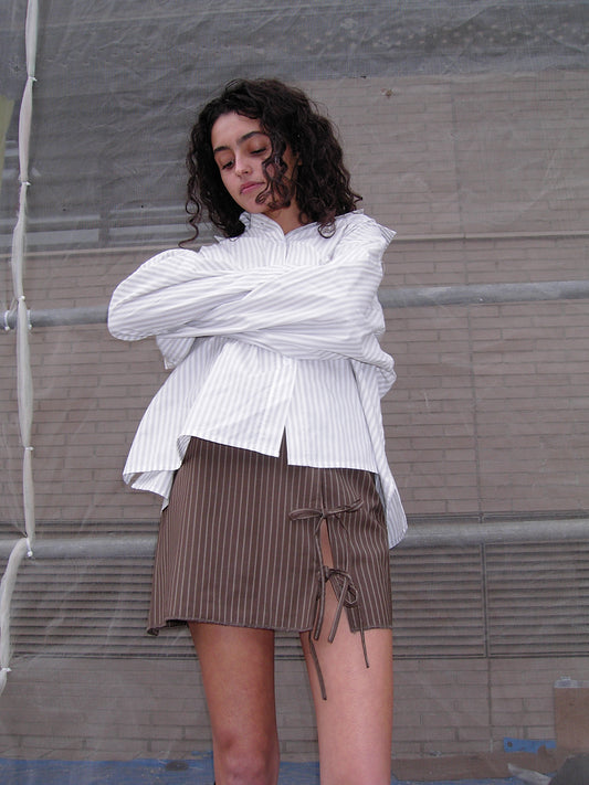 A person with curly hair stands with arms crossed, wearing a loose white striped shirt and the Two Ties Skirt—a brown pinstriped skirt with tie details—against a muted urban backdrop.