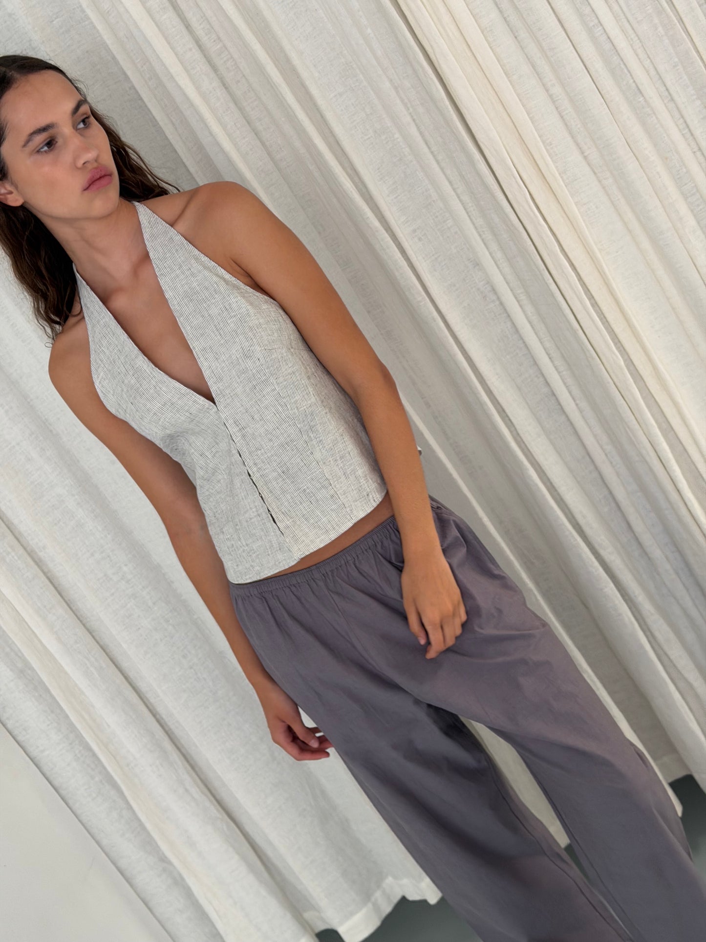 A woman with long brown hair stands in front of light curtains, wearing a sleeveless light grey halter top and Deiji Studios Ease Trouser in Currant. She gazes to the side with a relaxed expression.