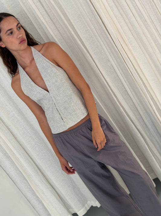 A woman with long brown hair stands in front of light curtains, wearing a sleeveless light grey halter top and Deiji Studios Ease Trouser in Currant. She gazes to the side with a relaxed expression.