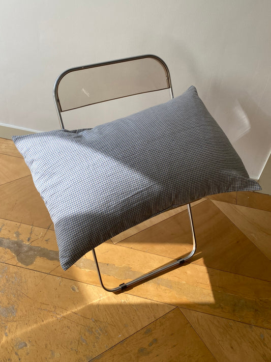 A large blue and white checkered pillow rests on a simple metal chair in a sunlit room with a wooden parquet floor. Sunlight casts shadows across the pillow and floor.