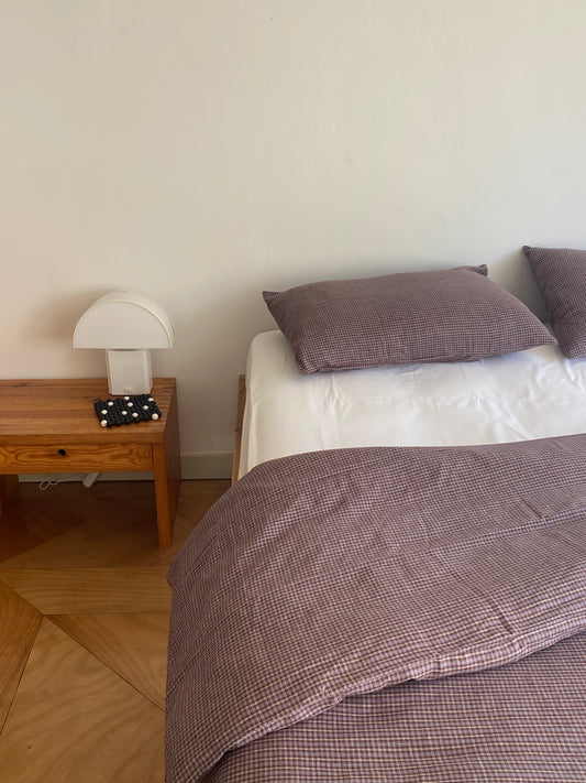 A neatly made bed with a Russet Check US linen duvet cover from Deiji Studios and two pillows sits beside a wooden nightstand holding a modern white lamp and black-and-white polka dot object, all set against a plain white wall.