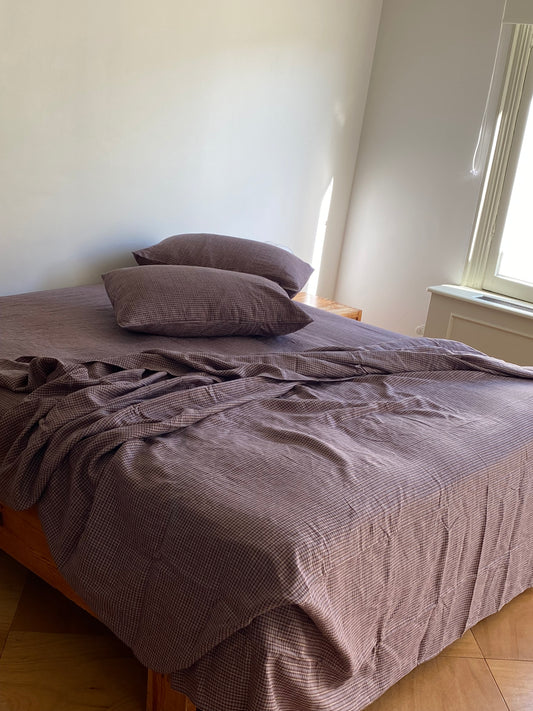 A wooden bed is dressed in Sheets - russet check with a brown checkered pattern and two stacked pillows. Sunlight from a right window highlights the slightly rumpled bedding.