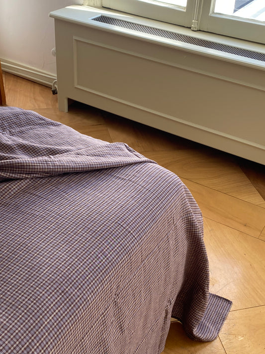 A hypo-allergenic blanket in purple and white check drapes over a bed with Sheets - russet check, set on wooden parquet flooring near a white radiator cover and a sunlit window.