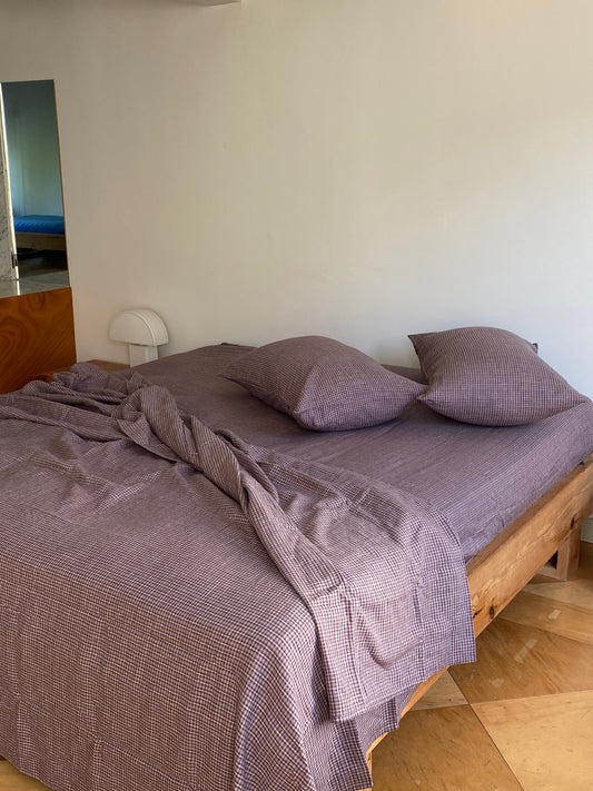 A wooden bed frame with breathable, wrinkled russet check linen sheets and two matching pillows, set on light hardwood floors by a white wall, with a mirror and small white lamp nearby.