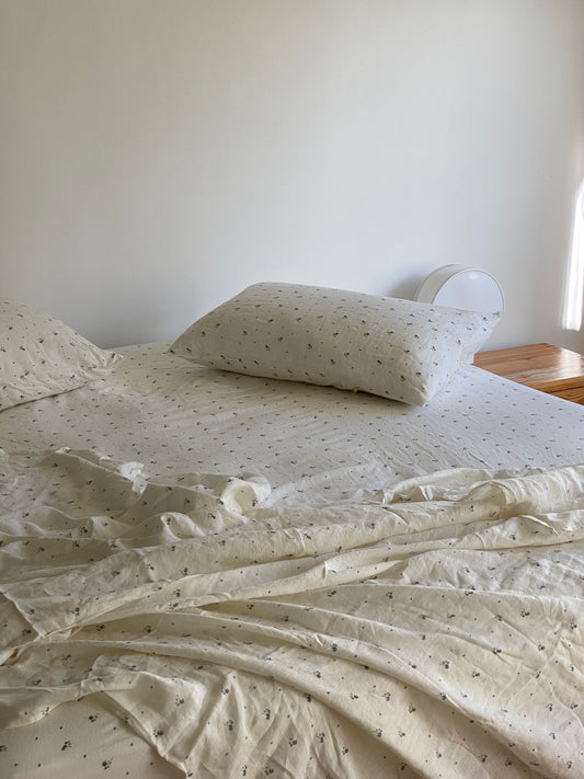 Unmade bed with vintage floral sheets and a pillow featuring a small black pattern. A wooden nightstand with a white round lamp sits beside the bed, sunlight streaming in from the right, against a plain white wall in the background.