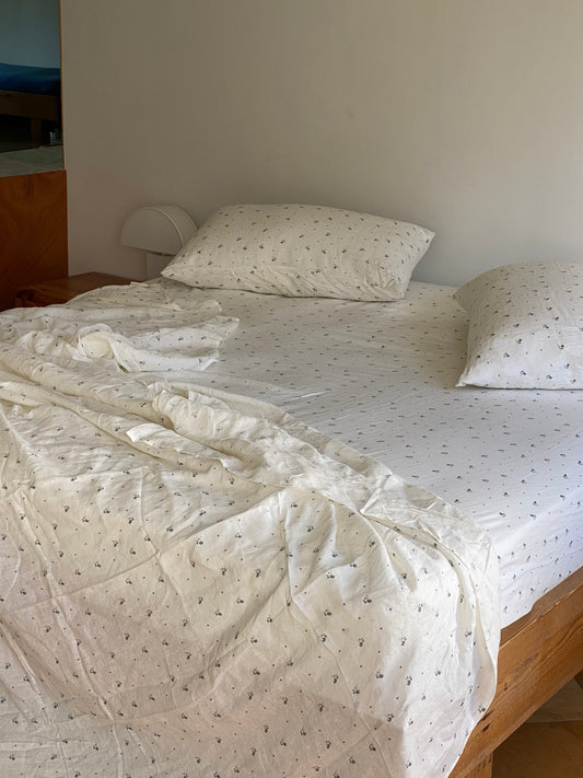 A bed with slightly rumpled Sheets - vintage floral and two pillows, both with a subtle pattern. A wooden nightstand with a small lamp is on the left side.