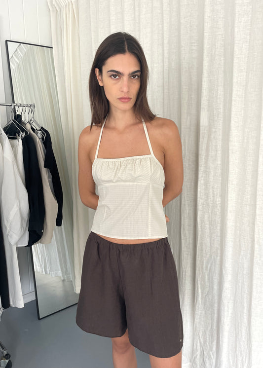 A woman with straight brown hair stands indoors against white curtains, wearing a white Strap Top and loose brown shorts, hands behind her back. Clothing racks and a mirror are in the background.