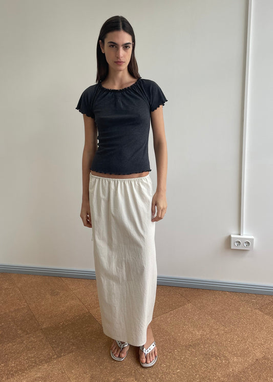 A woman with straight dark hair and a neutral expression stands indoors against a plain wall, wearing a black short-sleeve top, the Mid Day Skirt - Bone Check (a white A-line midi skirt with an elastic waistband), and white sandals.