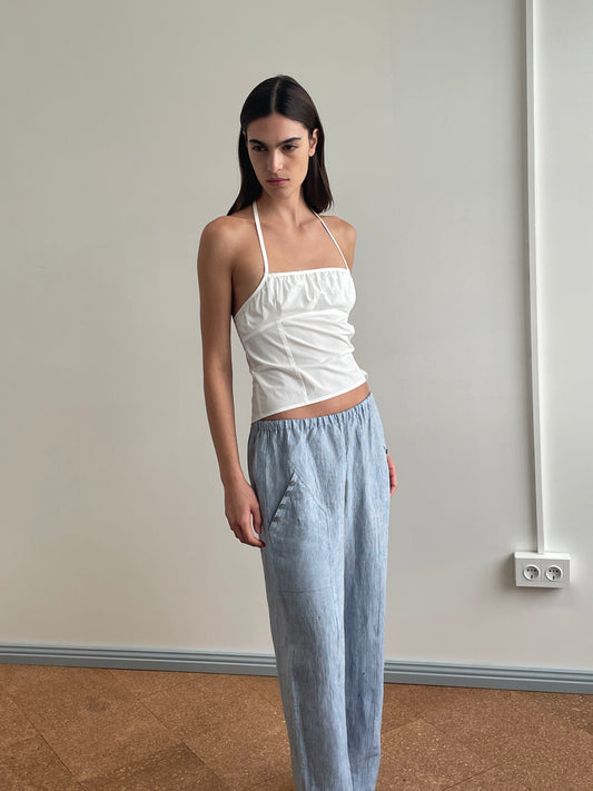 A woman stands indoors against a plain wall, wearing the Strap Top - White by Deiji Studios paired with loose-fitting light blue pants. Her hands are in her pockets, and she looks slightly to the side with a neutral expression.