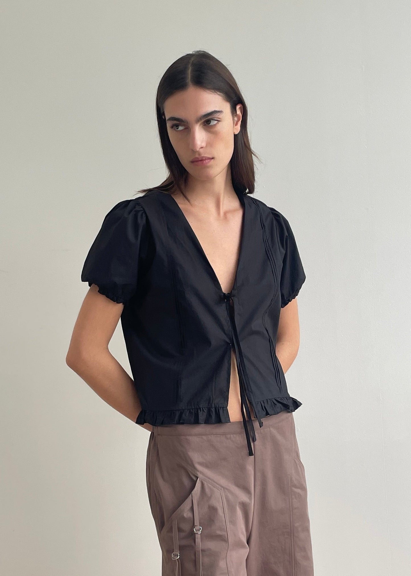 A person with straight dark hair wears the Pintuck Top - Black by Deiji Studios, featuring short sleeves and a deep V-neck, paired with brown cargo pants, standing against a plain light wall with hands behind their back.