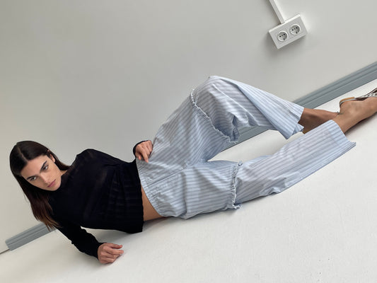 A woman with long dark hair lies on her side on a white floor, wearing a black long-sleeve top and Deiji Studios Flounce Short - Mineral Stripe, with a double electrical outlet visible on the wall behind her.