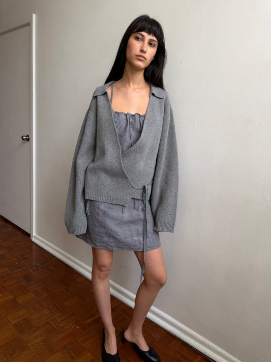 A woman with dark hair and a fringe stands against a wall wearing the Weave Mini - Indigo Check by Deiji Studios, an indigo micro-check linen mini dress, layered under a grey wrap cardigan with black flats.