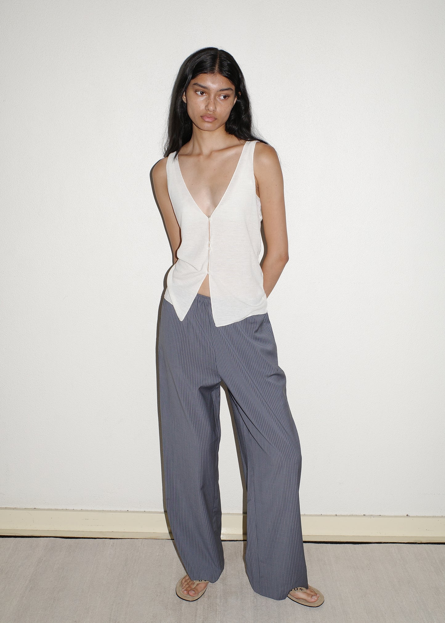 A woman with long dark hair stands against a plain wall, wearing the Button Up Knit Tank in Off White and loose-fitting, gray striped pants made from recycled polyester. She has her hands behind her back and is wearing sandals.
