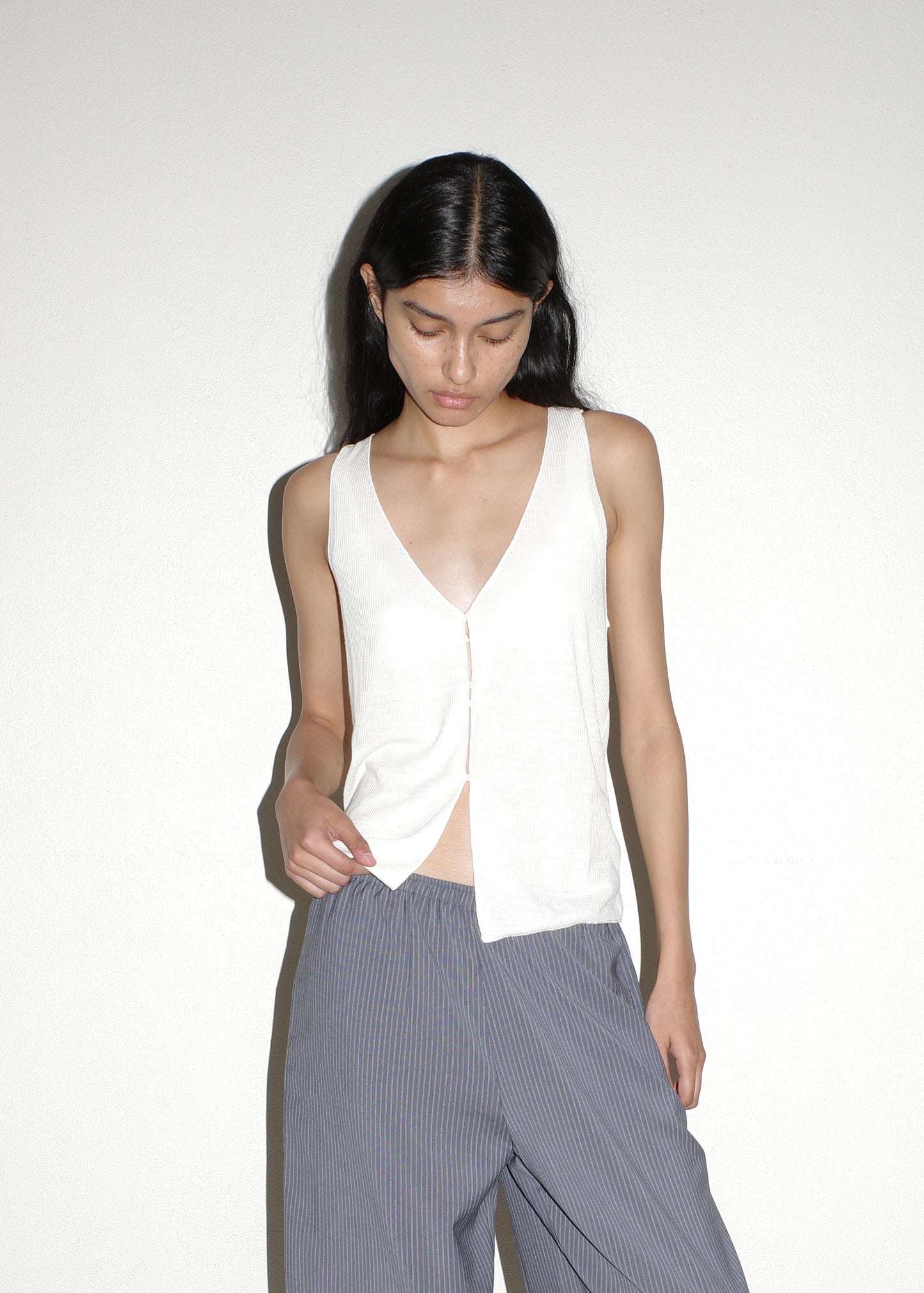 A young woman with long dark hair wears the Button Up Knit Tank in Off White, paired with loose gray striped pants, as she stands against a plain light-colored wall and looks down.