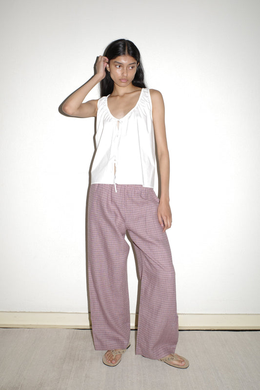 A woman with long dark hair stands against a white wall, wearing Linen Ease Trousers with a sleeveless white top and sandals. She touches her head with one hand and gazes off to the side.