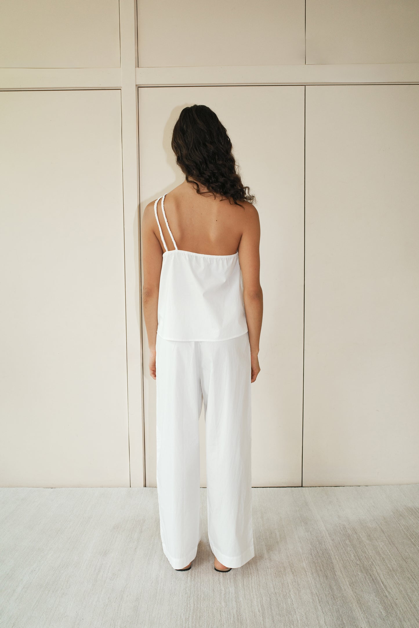 A woman with long dark hair stands barefoot facing a beige wall, wearing a Deiji Studios Shoulder Top - White with double straps and matching wide-leg pants. Her back is turned to the camera.