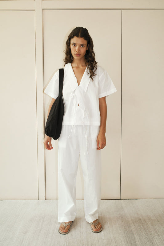 A person with long curly hair stands against a beige wall, wearing the Panel Shirt—a loose white short-sleeve top—with matching white pants, flip-flops, and carrying a black shoulder bag.