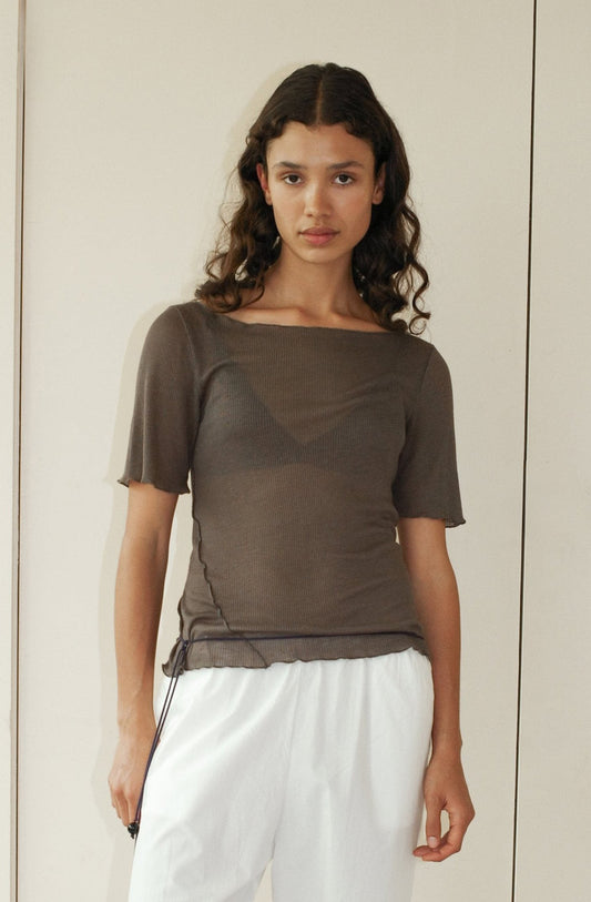 A woman with long, curly brown hair wears the Square Knit Tee - Stone by Deiji Studios over a bralette and loose white pants, standing against a plain, light-colored wall.