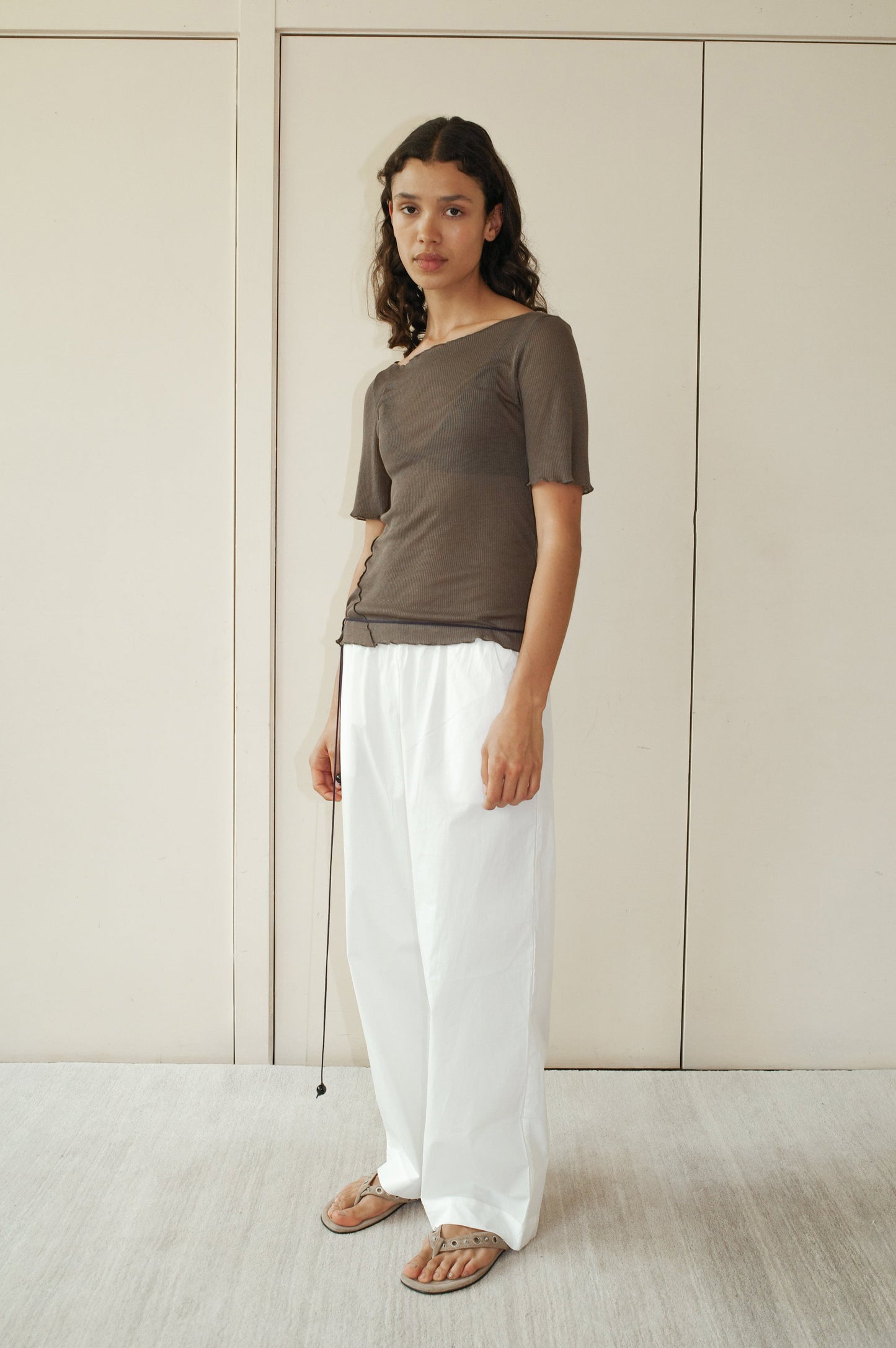 A person stands indoors wearing a Deiji Studios Square Knit Tee in Stone, loose white pants, and flat sandals against light-toned minimalistic wall panels.
