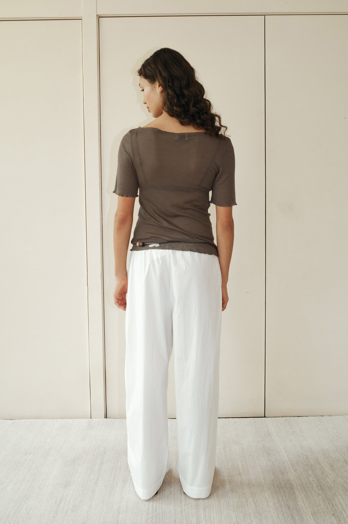 A woman with curly hair faces away from the camera, wearing Deiji Studios’ Square Knit Tee in Stone—a sheer brown top crafted from a responsible blend—paired with loose white pants, standing before beige paneled doors.