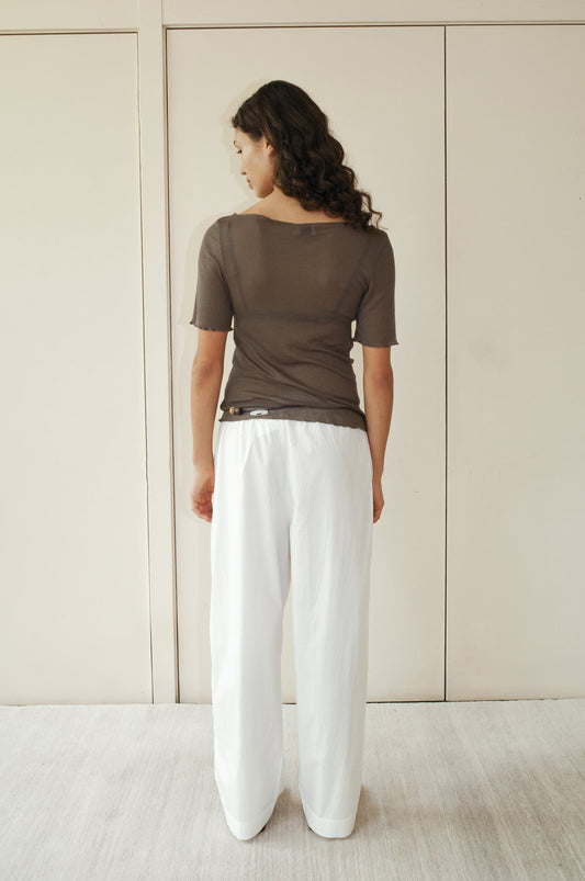 A woman with long wavy hair stands indoors, facing away from the camera in a Square Knit Tee - Stone by Deiji Studios and loose white pants, set against a light-colored wall.