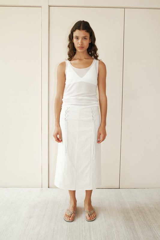 A woman stands indoors against a beige wall, wearing a white sleeveless top and the Double Lace Up Skirt - Bone Check with side ties. She pairs it with sandals, her long wavy hair flowing as she maintains a neutral expression.