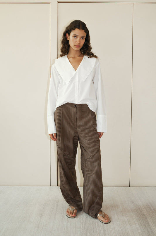 A woman stands indoors wearing a white long-sleeve blouse with a large collar, Channel Pant - Sable Stripe by Deiji Studios, and sandals. She has long wavy hair and a neutral expression, standing against beige-paneled doors.