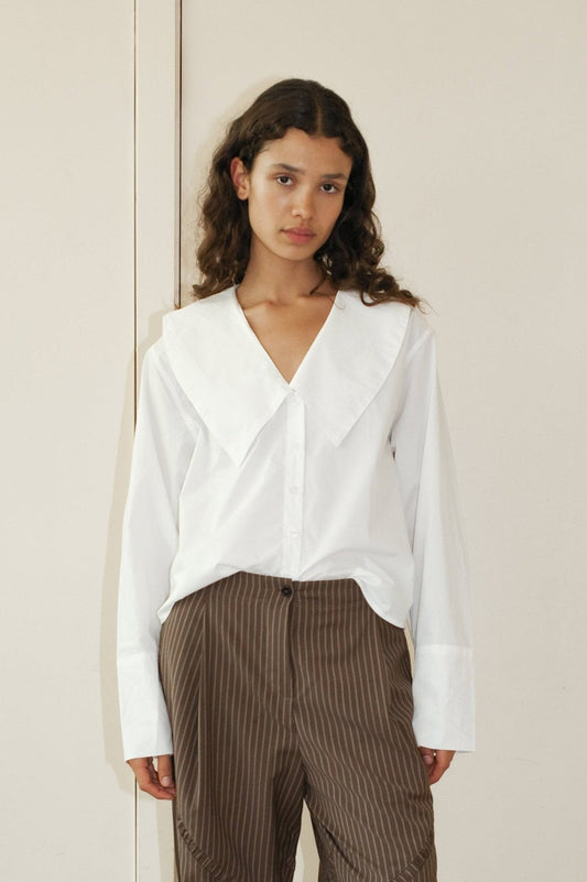 A young woman with wavy brown hair wears the Oversized Collared Shirt, featuring a large collar, paired with brown pinstripe trousers. She stands against a light wall and looks at the camera with a neutral expression.