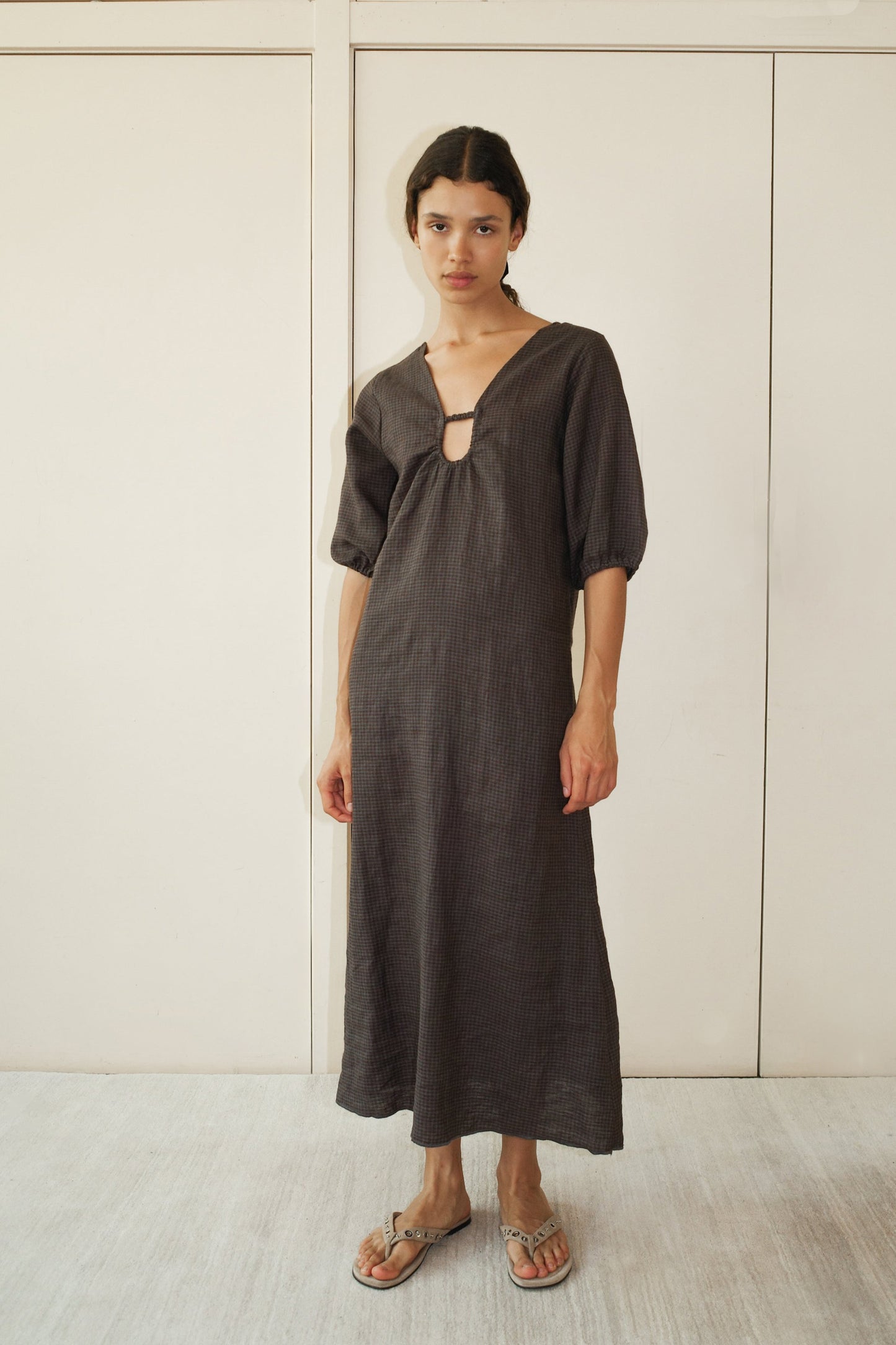 A woman stands indoors in the Squared Dress - Slate Check by Deiji Studios, an ankle-length, loose organic linen dress with elbow-length sleeves and a front keyhole neckline, paired with beige sandals against a light background.